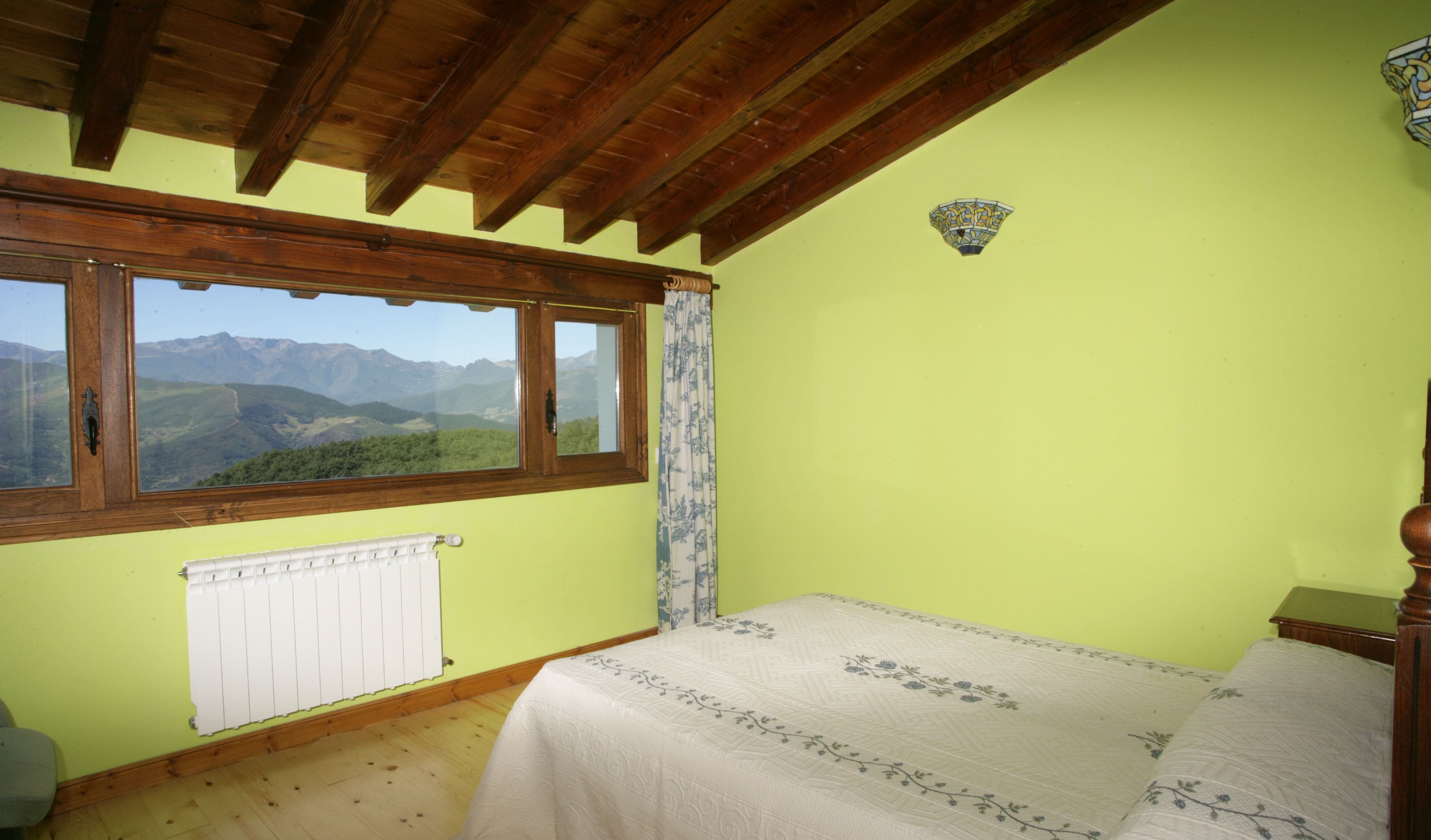 A bedroom with a bed, a radiator, a window with a view of mountains, and a wooden ceiling.