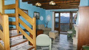 A cozy living area with a wooden staircase, a sofa, a chair, and a view of mountains through a balcony.