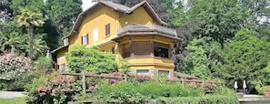 A yellow house with a balcony, surrounded by lush greenery and flowering plants.