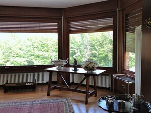 A room with a wooden table, a radiator, a window with blinds, and a view of greenery outside.