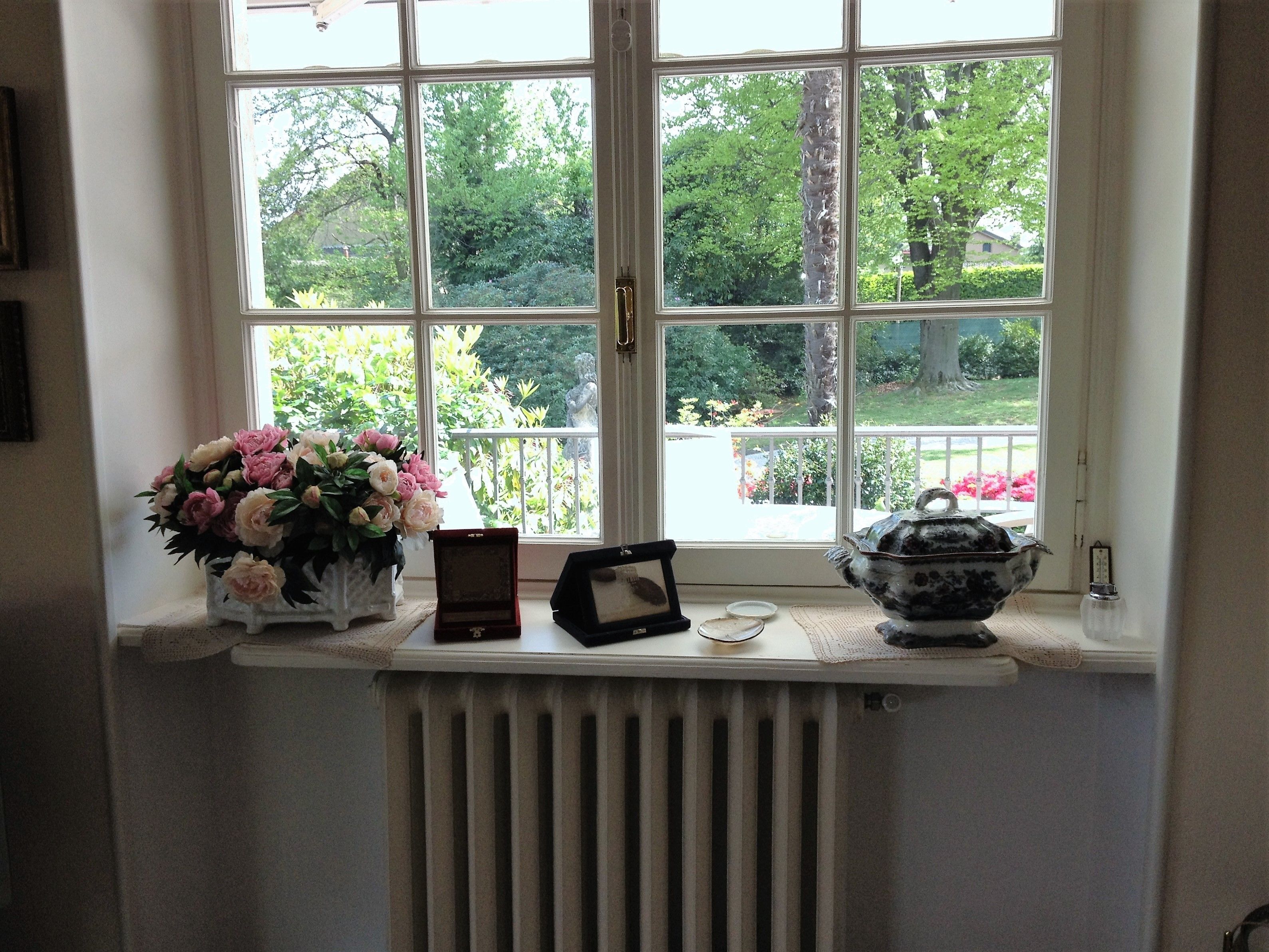 A window with a view of a garden, a vase of flowers, and a teapot on the windowsill.