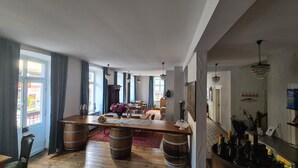 A spacious dining area with a long wooden table, multiple chairs, and a red rug. There are large windows with curtains, a chandelier, and a bar area with bottles. - Osteria Del Vino Cochem (Cochem)