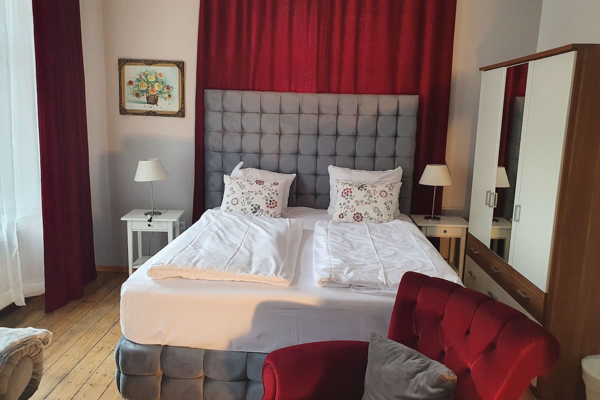 Uma cama bem arrumada com lençóis brancos e almofadas florais, uma poltrona estofada em vermelho, uma mesinha de cabeceira com abajur e um quadro na parede.