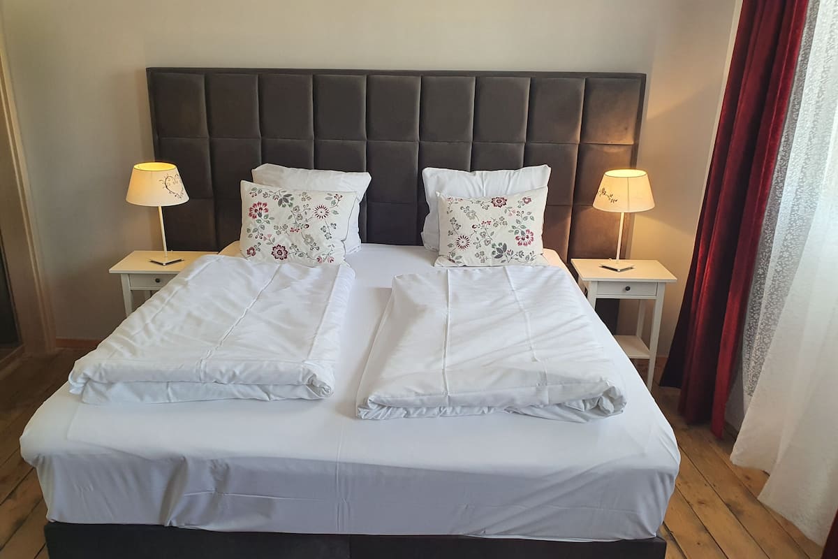 Cama bem arrumada com lençóis brancos, dois travesseiros decorativos com estampas florais e duas luminárias de cabeceira sobre mesas de cabeceira combinando.