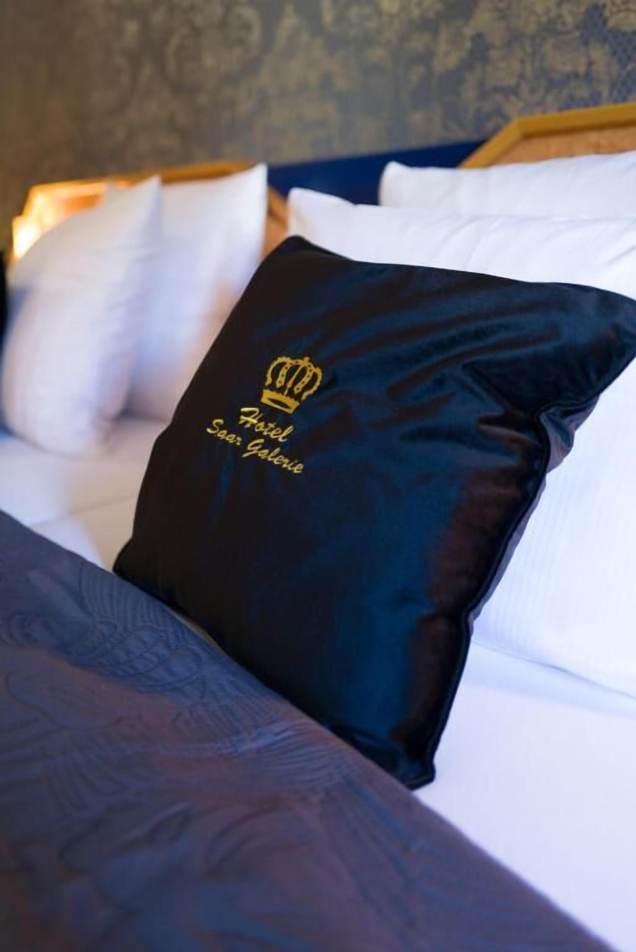 Una cama con un almohadón azul oscuro con una corona y texto, un almohadón blanco y una colcha estampada.