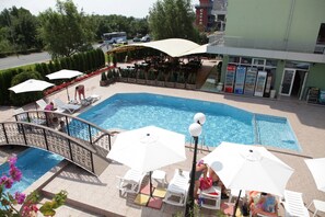 Una piscina con sillones de descanso y sombrillas, rodeada por un restaurante y un edificio con máquinas expendedoras.