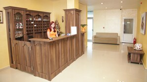 Un mostrador de recepción con detalles de madera, una recepcionista y una zona de espera con un sofá y una pequeña mesa con un florero.