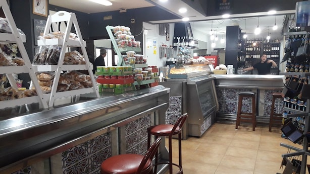 Mostrador de panadería con pasteles y pan, vitrina refrigerada y barra con taburetes.