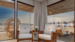 A room with large windows, wicker chairs, a wooden table with a vase of flowers, and a view of beach huts. - Regency Sealine Camp (Mesaieed)