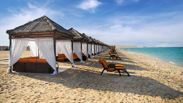 A beach with thatched-roof cabanas and orange lounge chairs. - Regency Sealine Camp (Mesaieed)