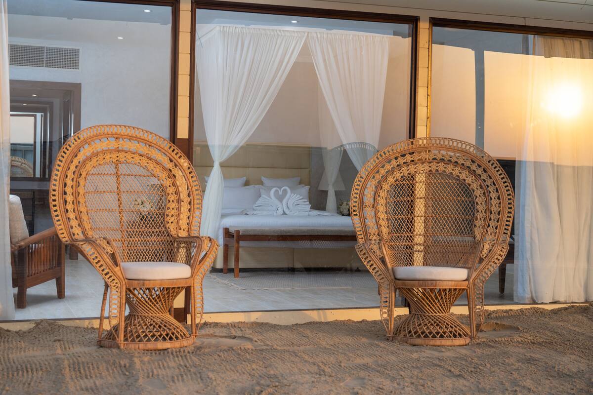 Two wicker chairs with cushions in front of a bed with white bedding and a canopy.