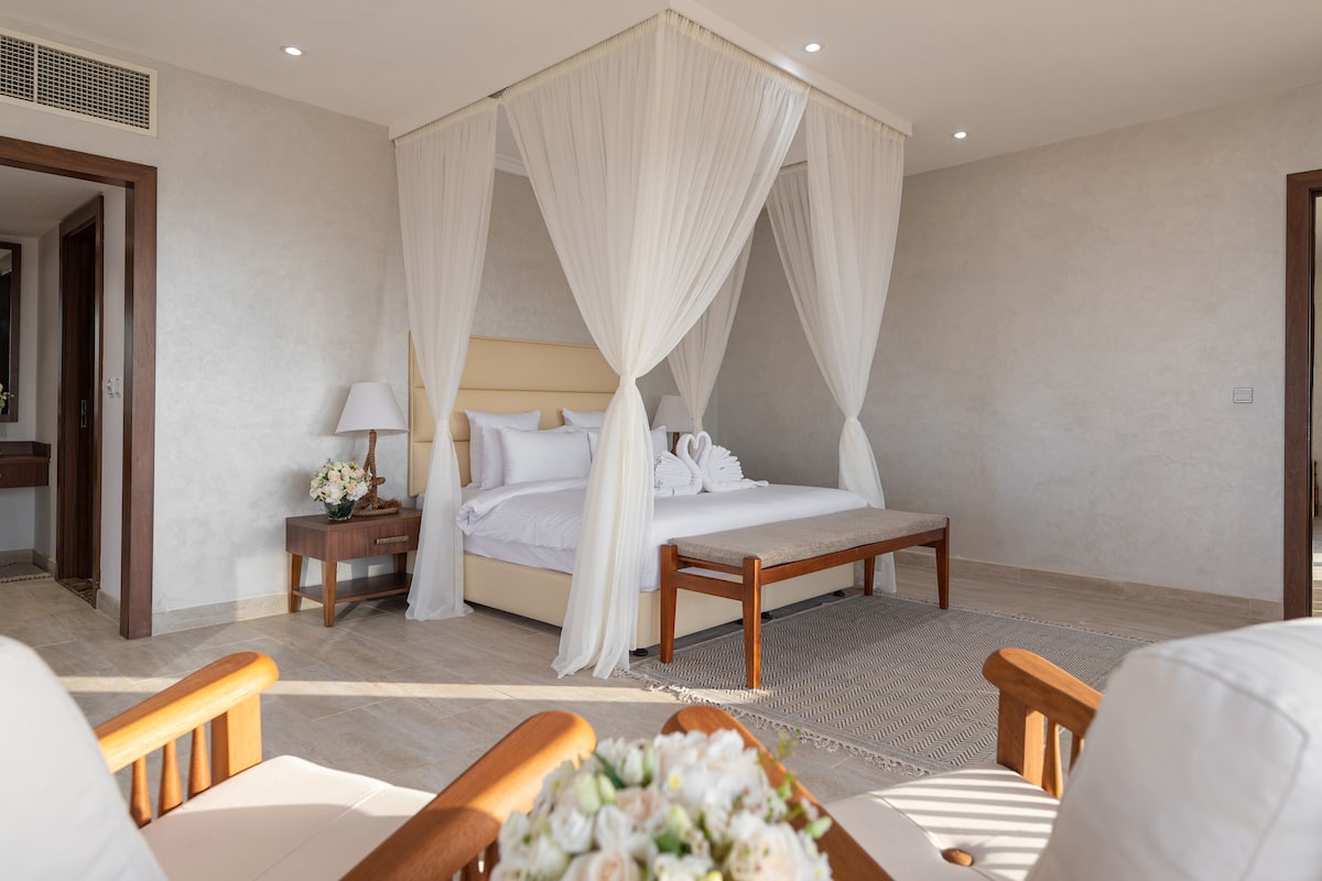 A bedroom with a four-poster bed, a bedside table with a lamp, a bench, and a chair.
