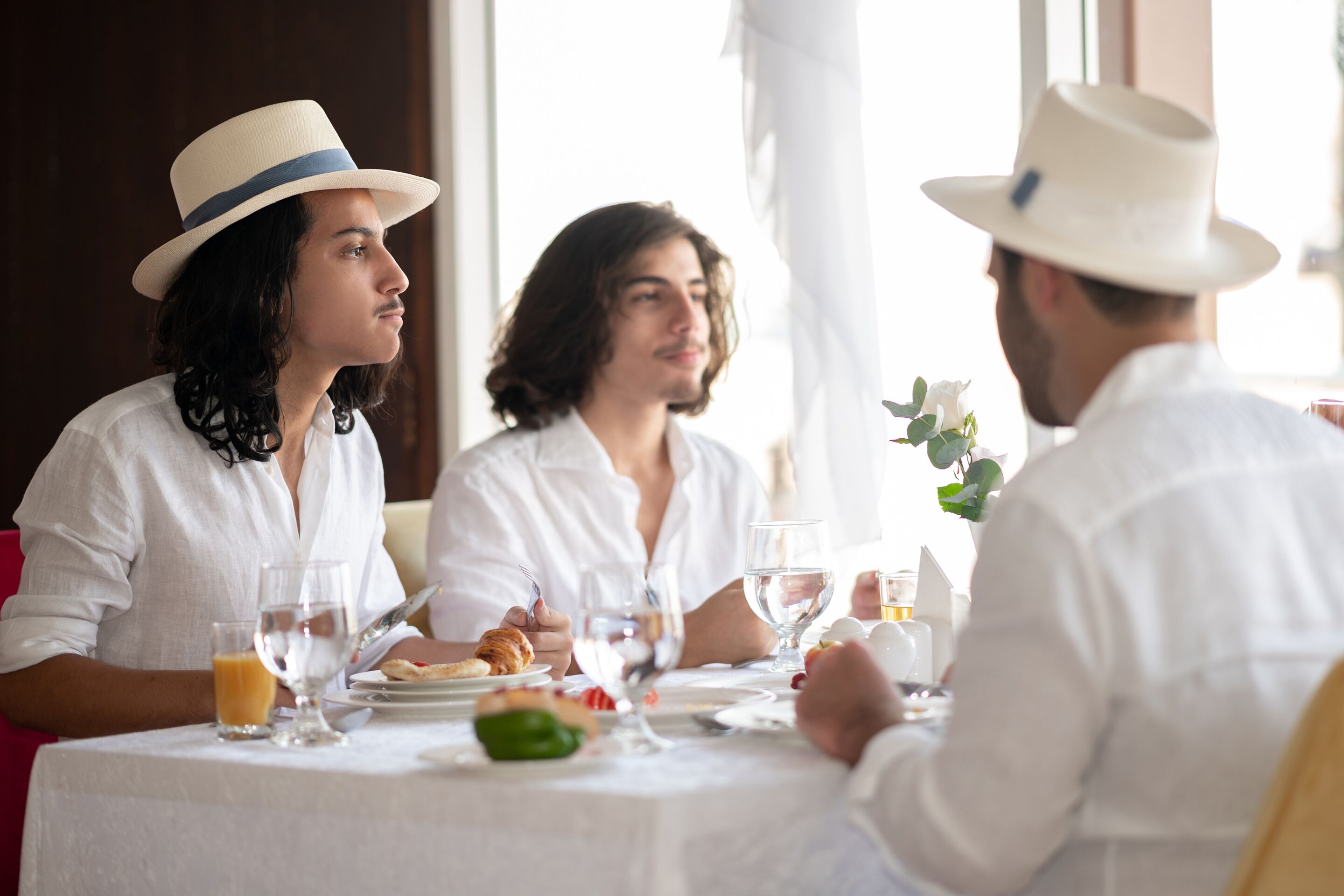 Uma mesa de jantar com toalha branca, pratos de comida, copos de água e suco, e um arranjo floral branco no centro.