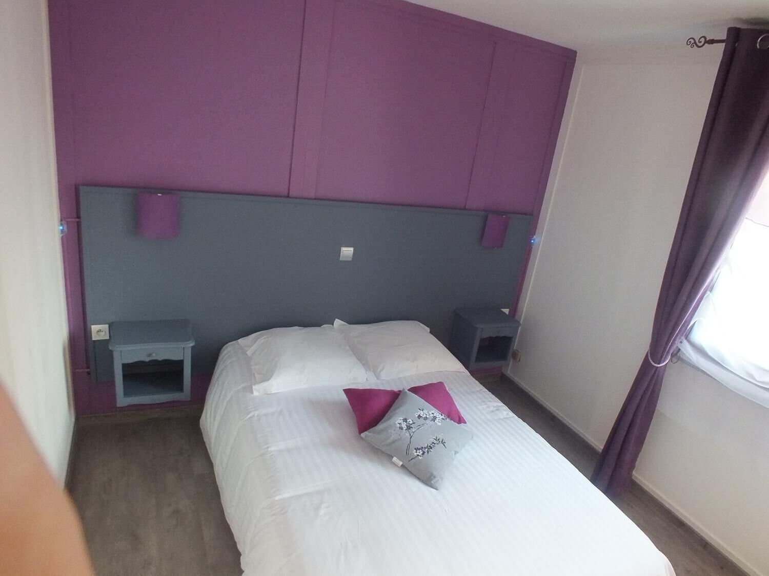 A small, neatly made bed with white linens and two pillows, one with a floral design. There are two bedside tables, one on each side of the bed. The room has a purple headboard and a window with curtains.