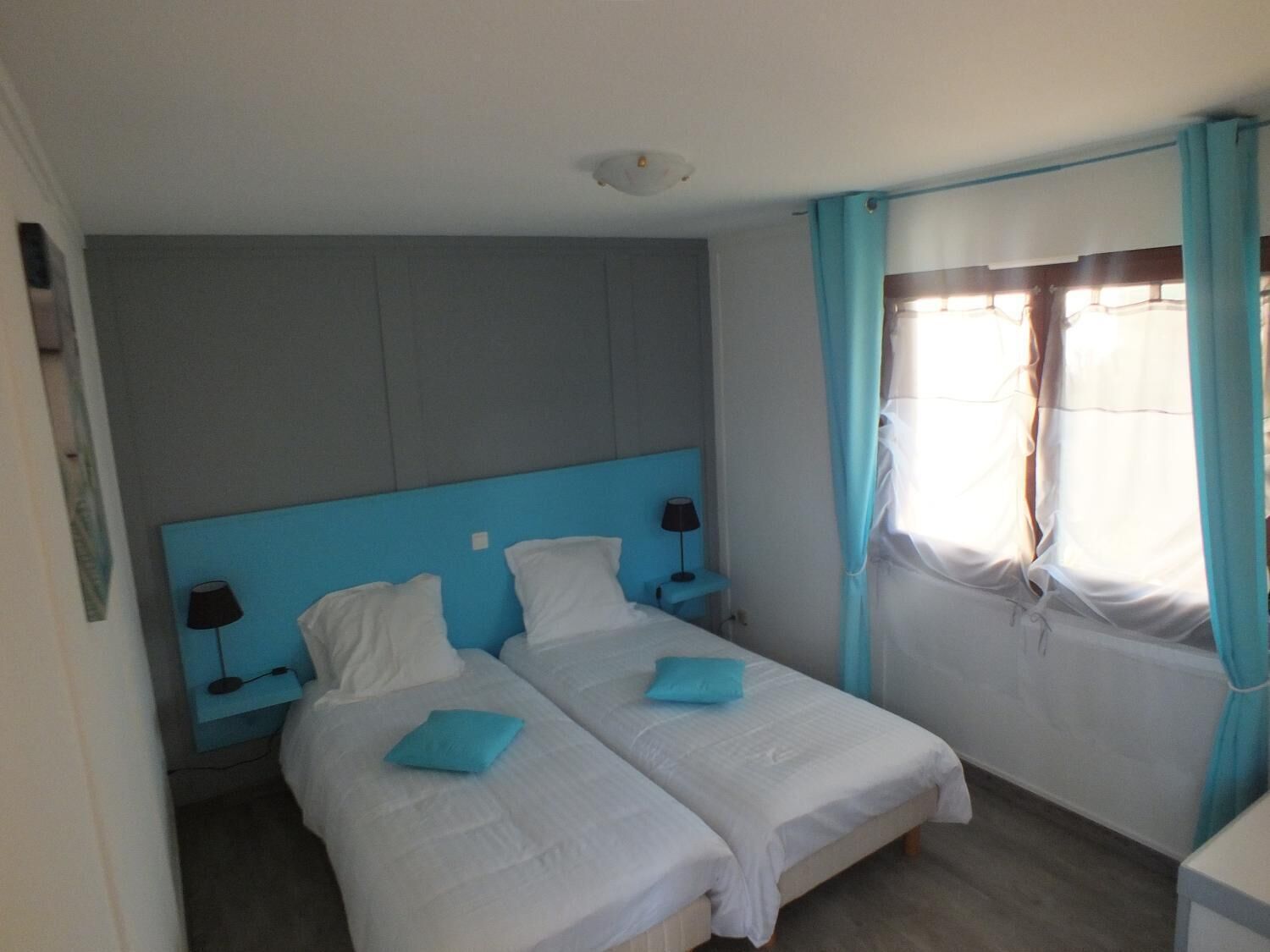 A hotel room with two beds, a blue headboard, white bedding, a nightstand with a lamp, and a window with curtains.