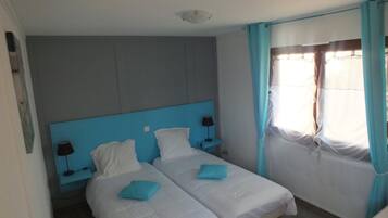 A hotel room with two beds, a blue headboard, white bedding, a nightstand with a lamp, and a window with curtains.