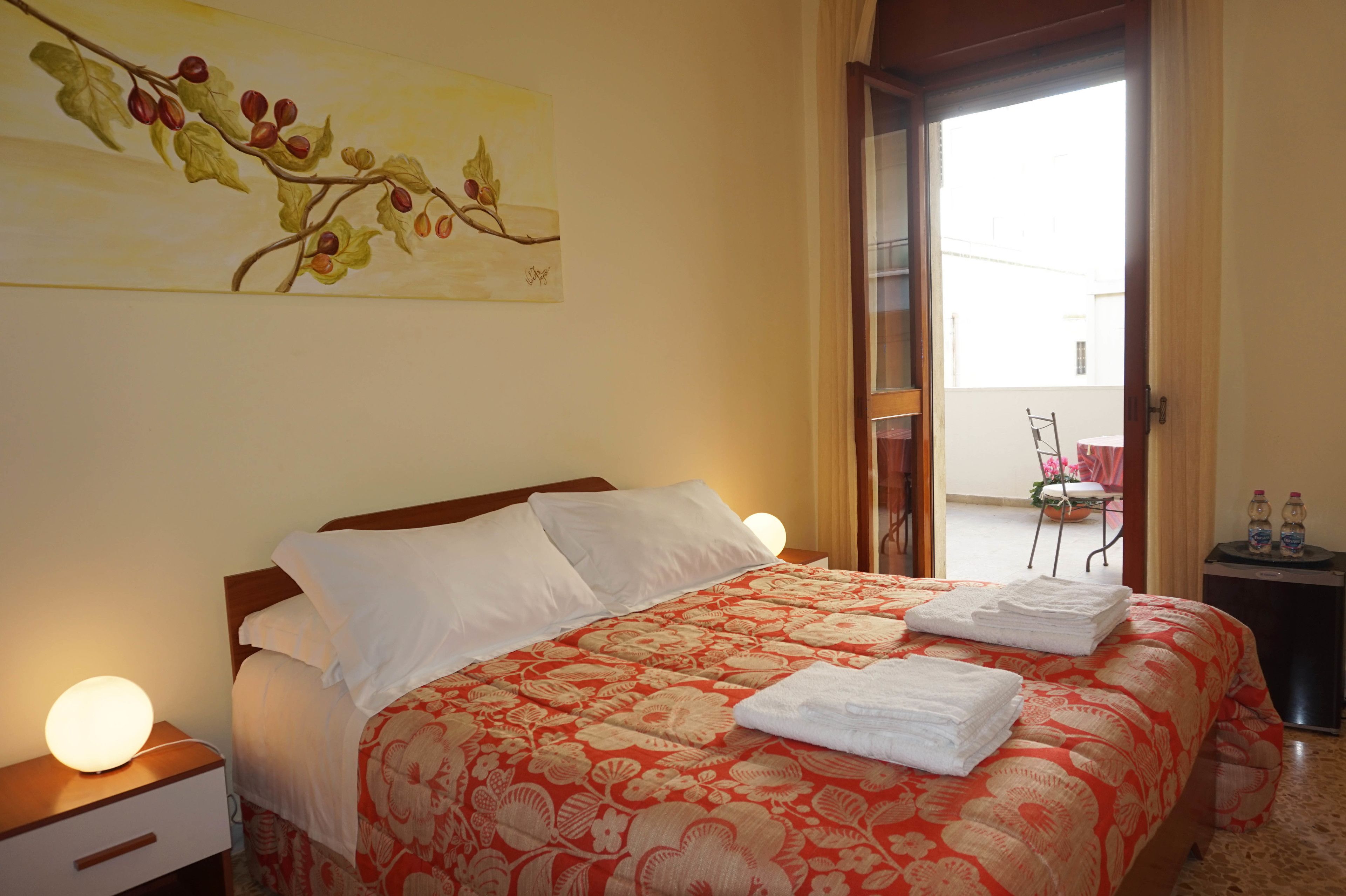 A hotel room with a bed, a nightstand, a painting, a balcony, and a small table with bottles.