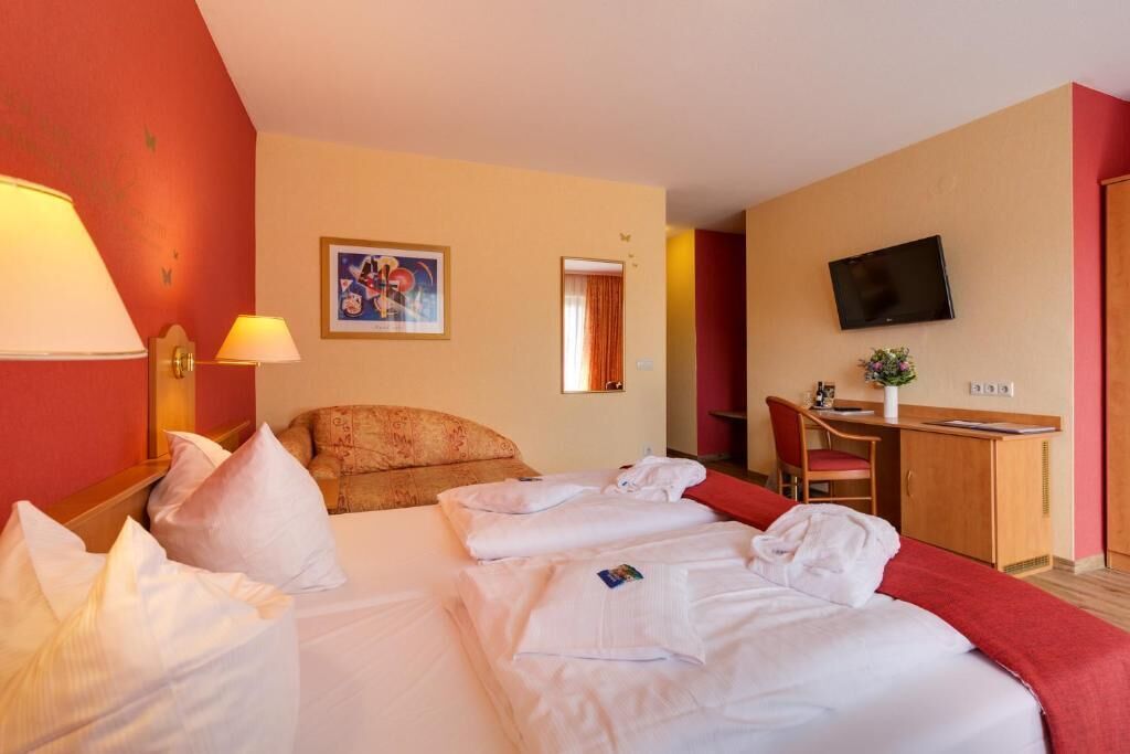 A hotel room with a bed, a desk, a chair, a TV, and a painting on the wall.