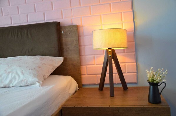 Una cama con cabecera, una lámpara de noche sobre una mesita de madera y una pequeña planta en maceta.