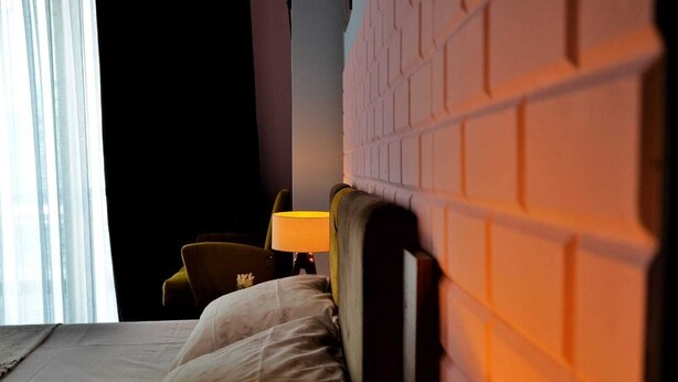 A hotel room with a brick-patterned headboard, a bed with a white bedspread, two lamps with yellow shades, and a chair with a floral cushion. - Retro Suites (Istanbul)