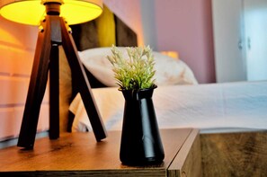 A bedside table with a black vase containing a plant, a wooden nightstand, and a lamp with a yellow shade.