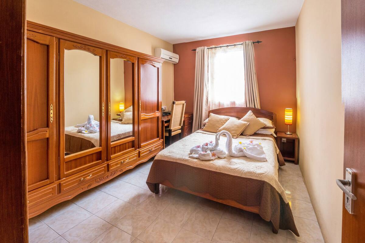 Una habitación de hotel con cama, armario, mesita de noche con lámpara y ventana con cortinas.
