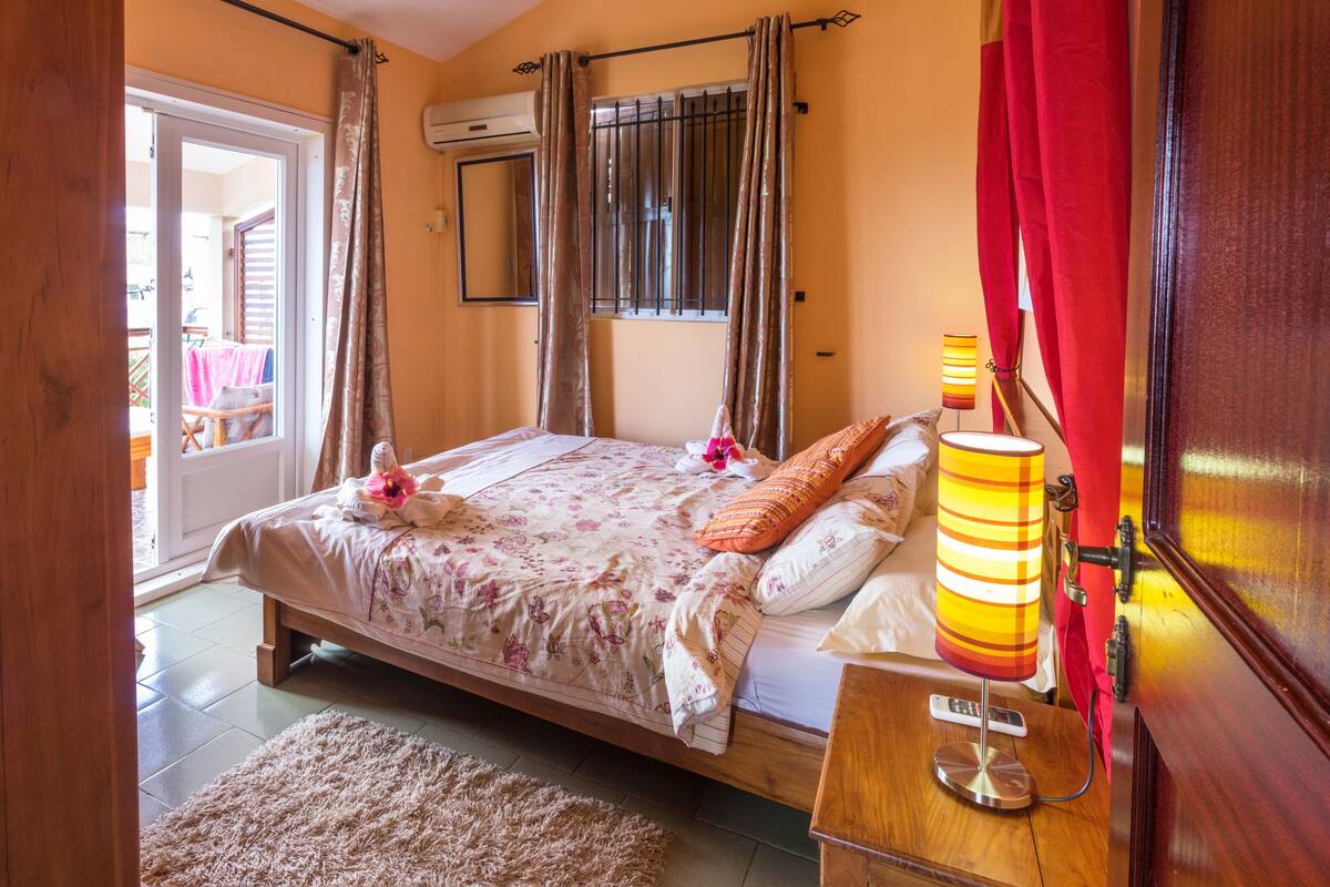 Un dormitorio con cama, una mesita de noche con lámpara, cabecero de madera, ventana con cortinas y vista al balcón exterior.