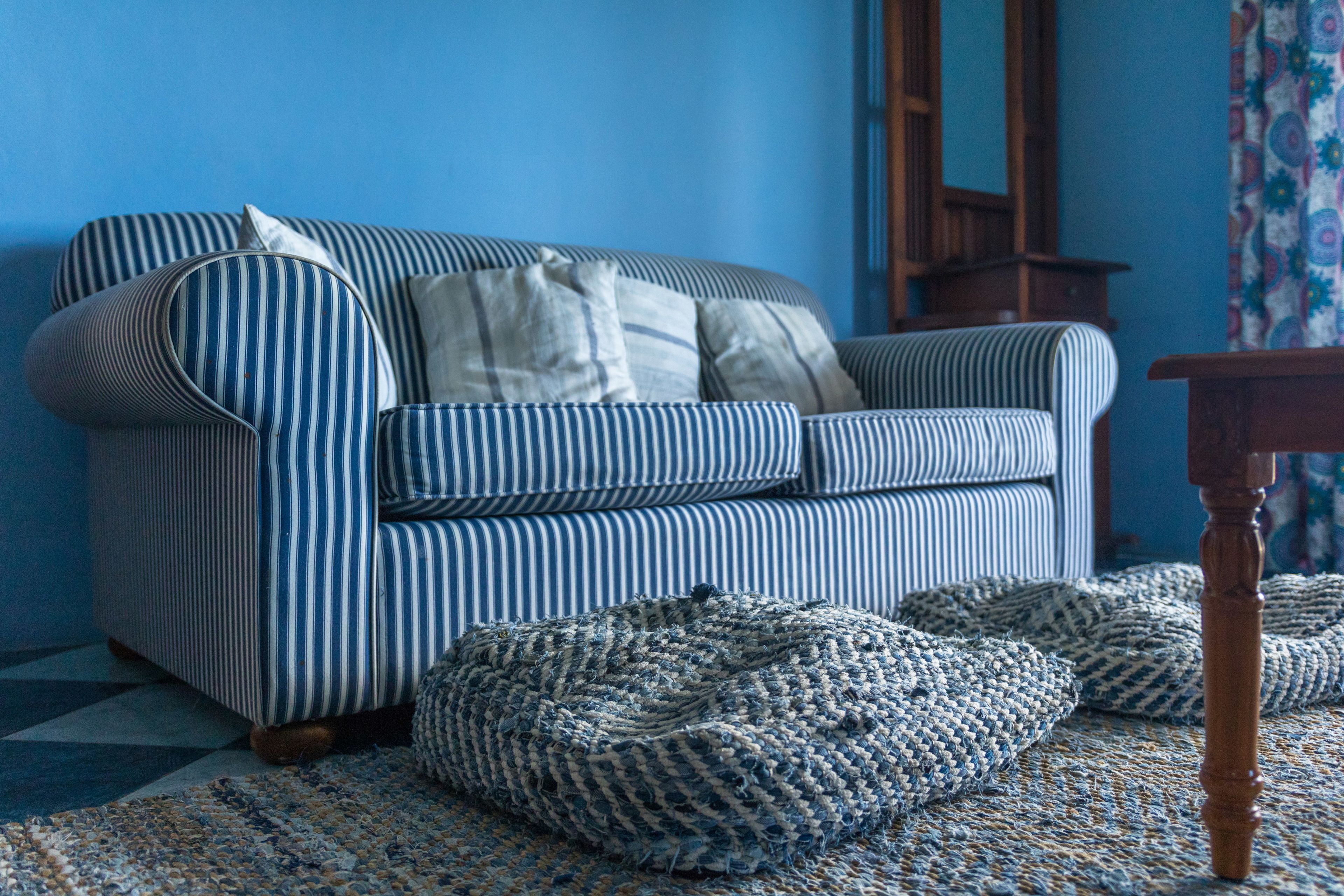 Un sofá a rayas con varios cojines, un taburete tejido y una mesa de madera en una habitación con paredes azules.