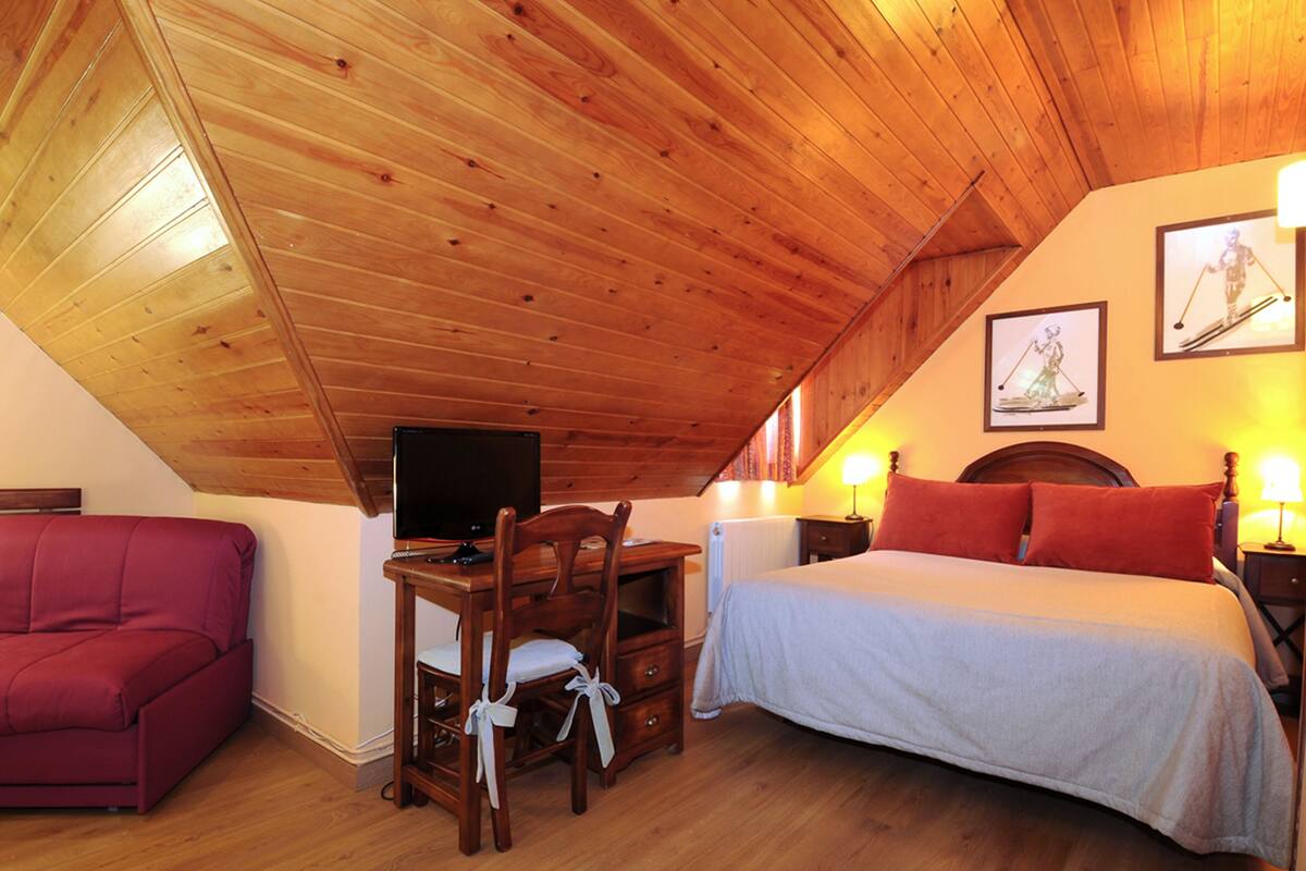 Ein gemütliches Zimmer mit schrägverlaufender Holzdecke, einem Bett mit roten Kissen, einem Schreibtisch mit Stuhl und einem Fernseher.
