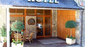 L’entrée d’un hôtel, avec une enseigne indiquant « HÔTEL » au-dessus de la porte, une façade en pierre et un petit espace extérieur aménagé avec des chaises et des plantes en pot.