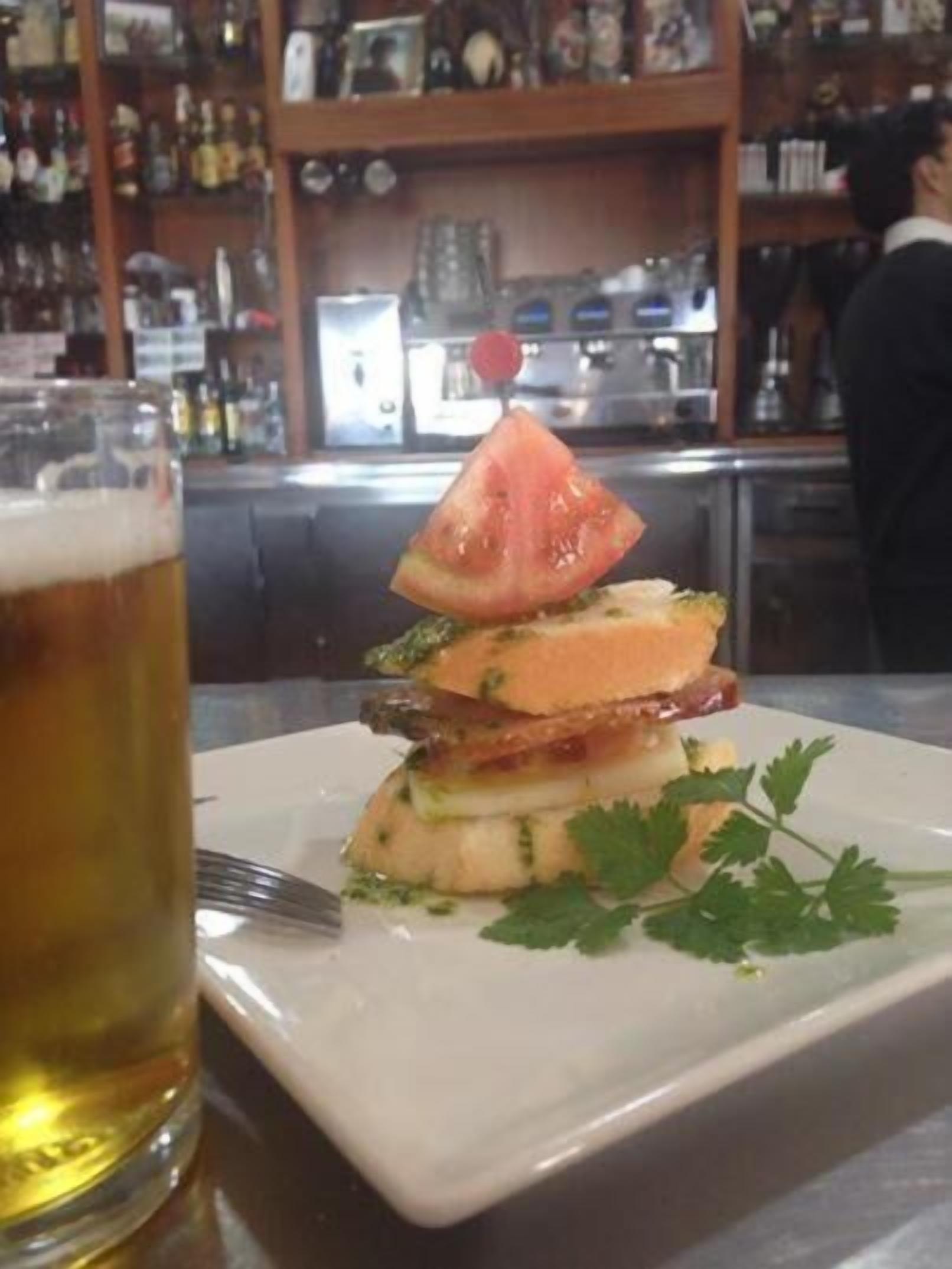 Un vaso de cerveza sobre una mesa con un plato de comida que incluye un sándwich, tomate y perejil.