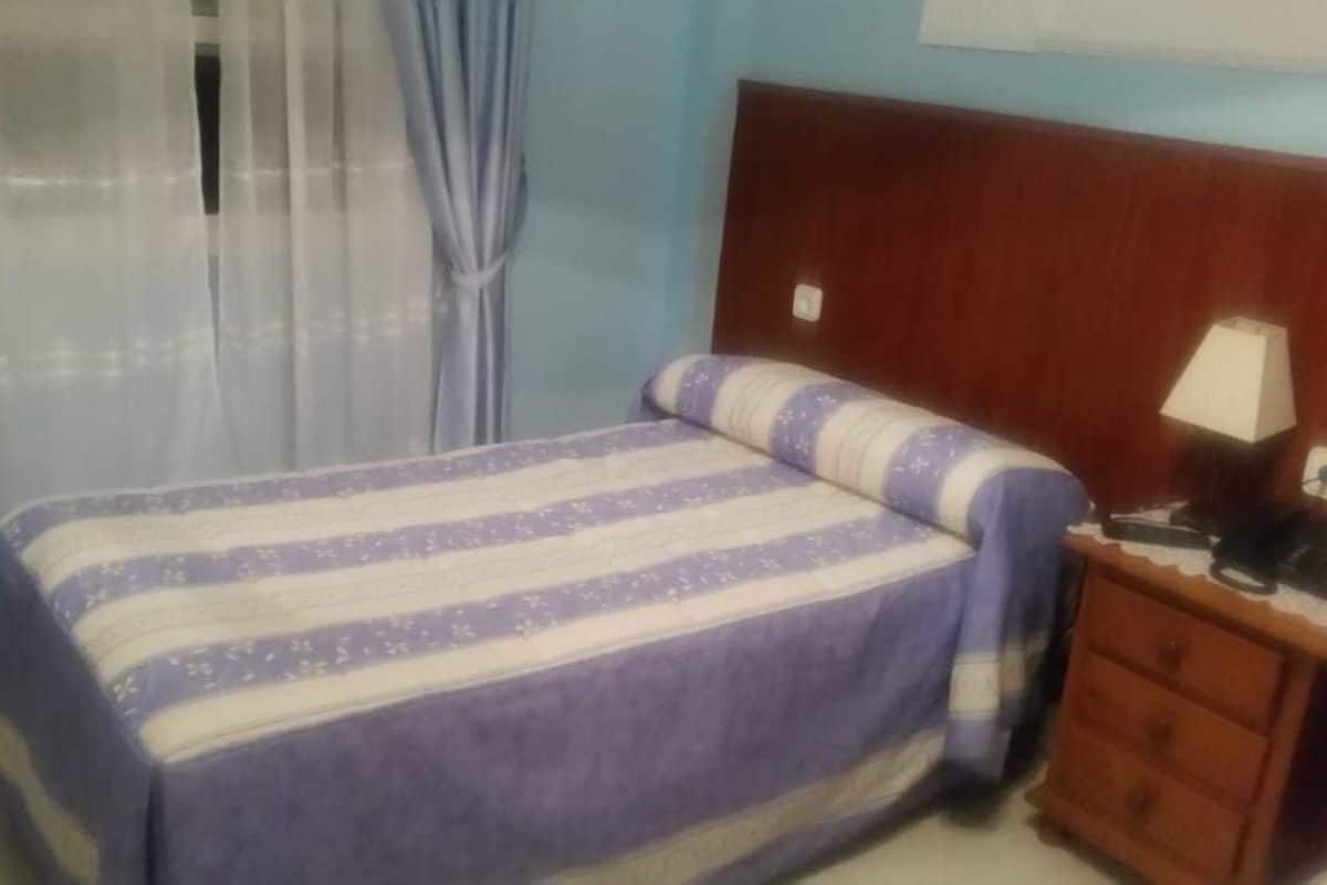 Een slaapkamer met een bed, een houten nachtkastje, een lamp en een raam met gordijnen.
