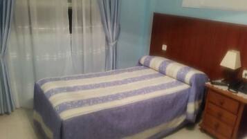 Un dormitorio con una cama, una mesita de noche de madera, una lámpara y una ventana con cortinas.