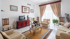 Un salón con sofá, sillones, una mesa de centro y televisor. Hay cuadros enmarcados en la pared y una ventana con cortinas.