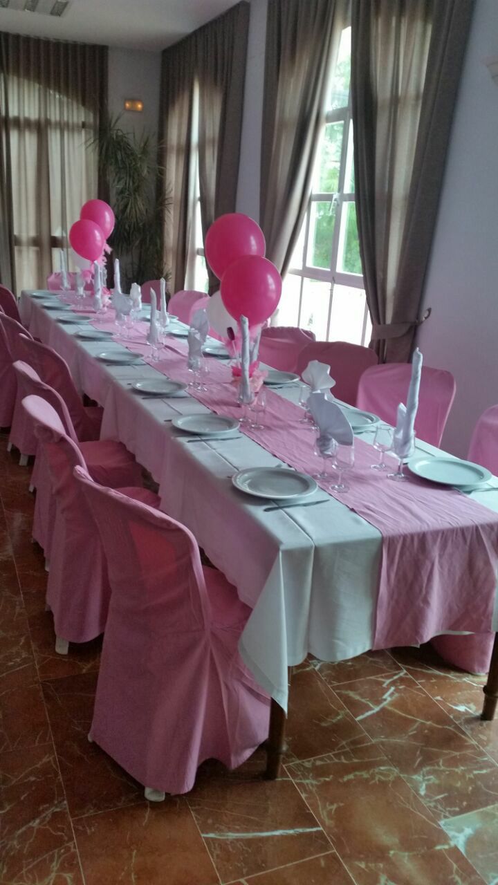 Une longue table à manger, entourée de chaises roses, dressée avec de l’argenterie, de l’assiette blanche et des ballons roses.
