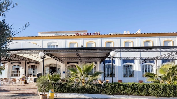 Un hotel con una zona de estar al aire libre techada, palmeras y un cielo azul despejado.