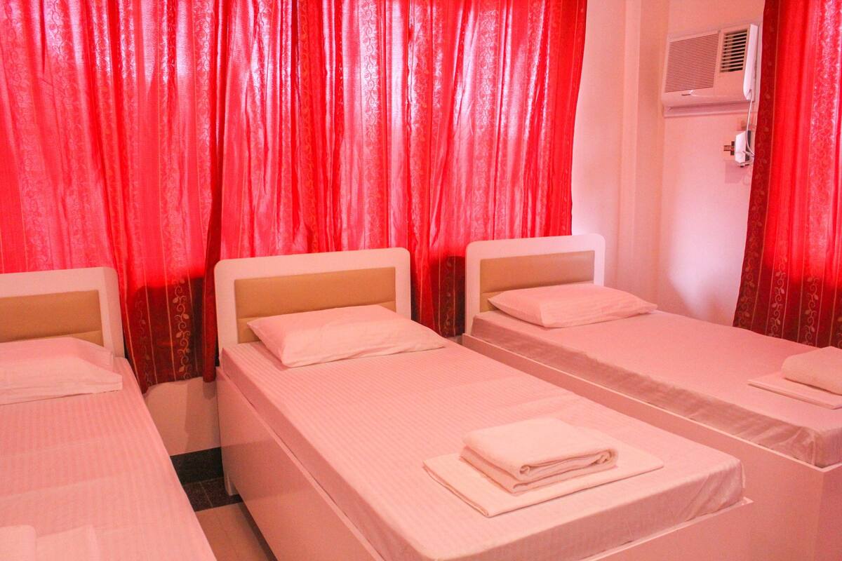 Ein Hotelzimmer mit drei Einzelbetten, jedes mit einem weißen Kopfteil und einem ordentlich gefalteten weißen Handtuch.