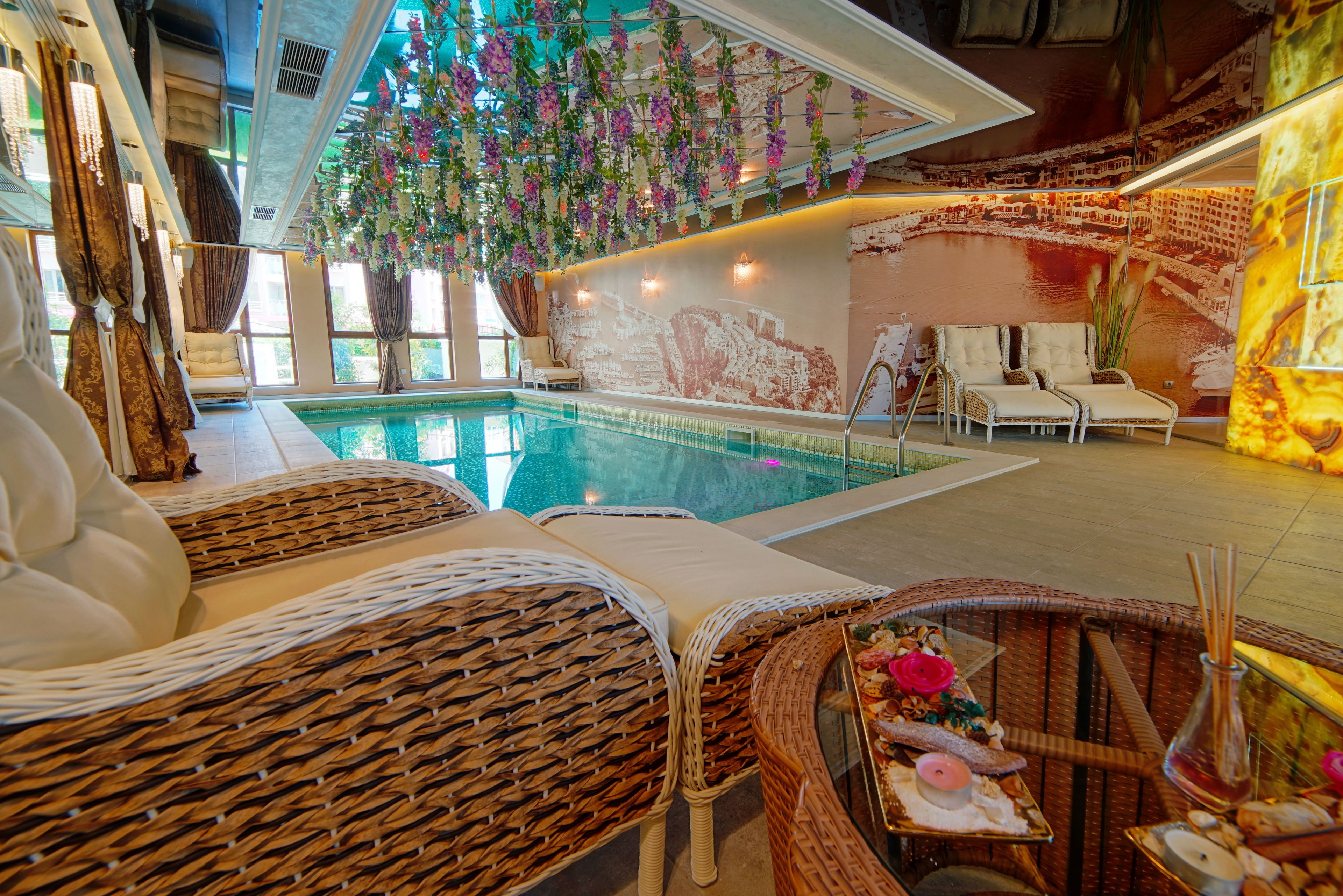 Uma área de piscina com espreguiçadeiras de vime, uma mesa com petiscos e uma parede decorativa com flores pendentes.