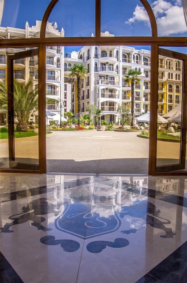 A hotel entrance with a reflective floor, a large arched doorway, and a view of multi-story buildings with balconies and palm trees. - Harmony Suites Monte Carlo (Sunny Beach)