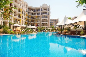 A large swimming pool with lounge chairs and umbrellas, surrounded by multi-story buildings with balconies.