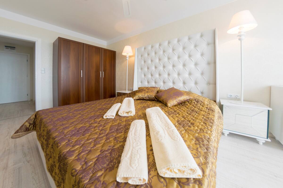 A neatly made bed with a patterned bedspread and two rolled towels, a white tufted headboard, a bedside table with a lamp, and a wooden wardrobe.