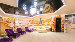 Un hall d’entrée spacieux avec un plafond haut, un grand lustre et un coin salon agrémenté de fauteuils violets et d’un canapé blanc.