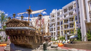 Un espace aquatique sur le thème des pirates, avec un navire et un canon, entouré d’une piscine et d’un espace de détente, et un immeuble d’hôtel à plusieurs étages en arrière-plan.
