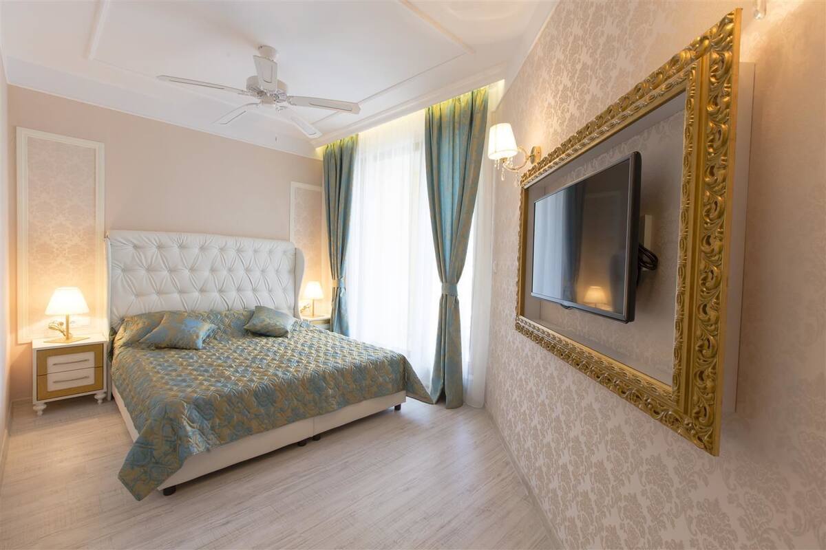 A bedroom with a tufted headboard, a bed with patterned bedding, a television mounted on the wall, and a ceiling fan.