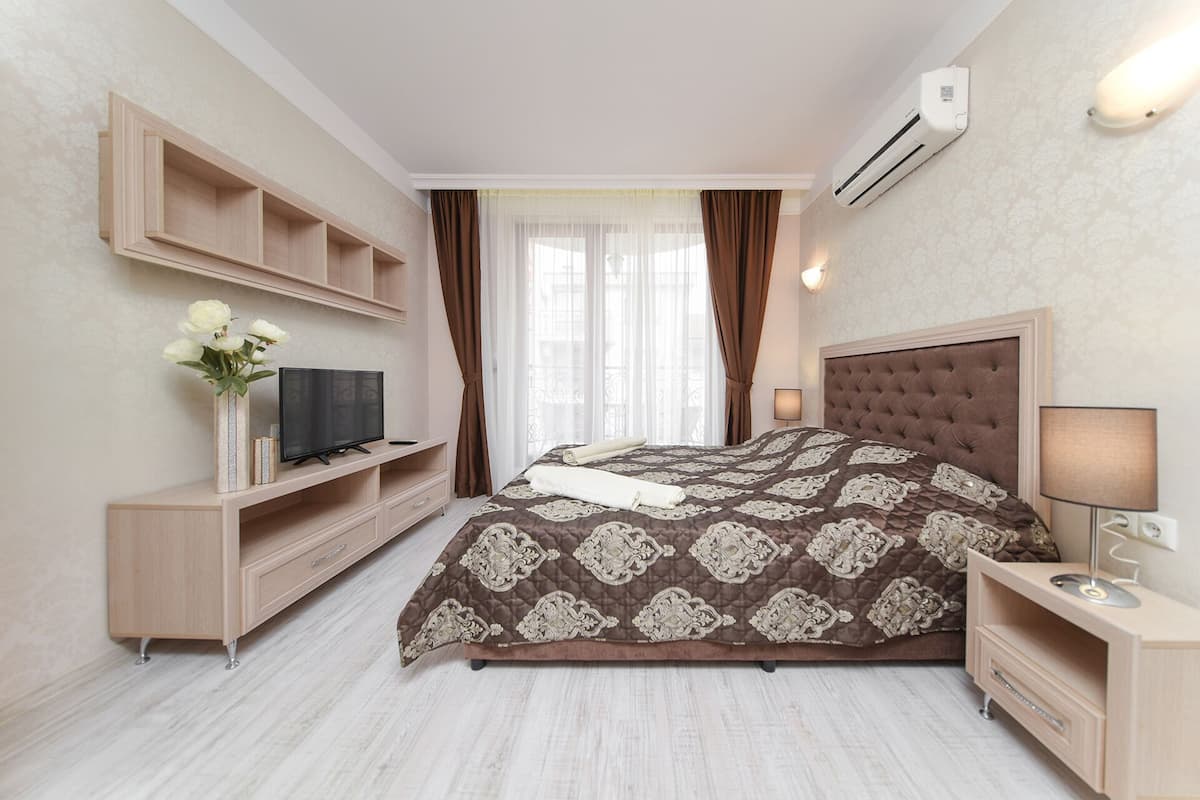 A bedroom with a large bed, a TV on a wooden stand, a bedside table with a lamp, and an air conditioning unit on the wall.