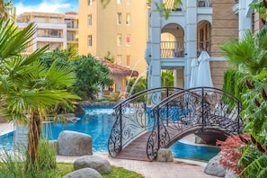 Area piscina con un ponte decorativo, circondata da piante tropicali e un edificio a più piani sullo sfondo.