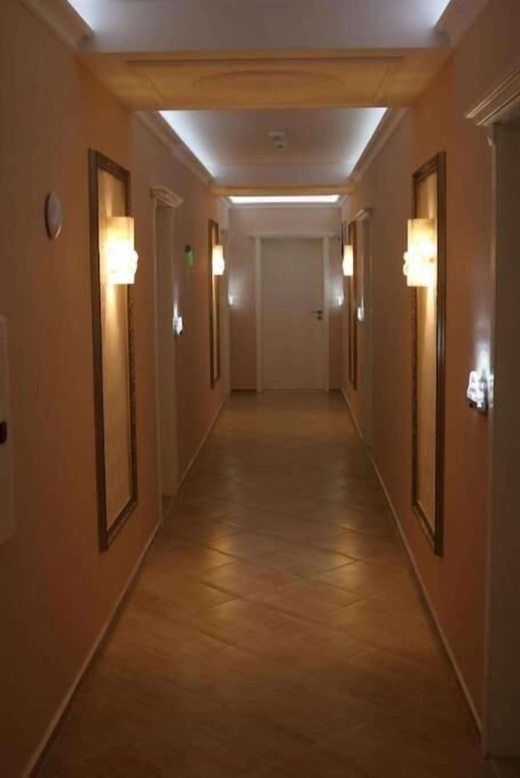 A hotel hallway with wooden floors, light-colored walls, and wall-mounted lamps.