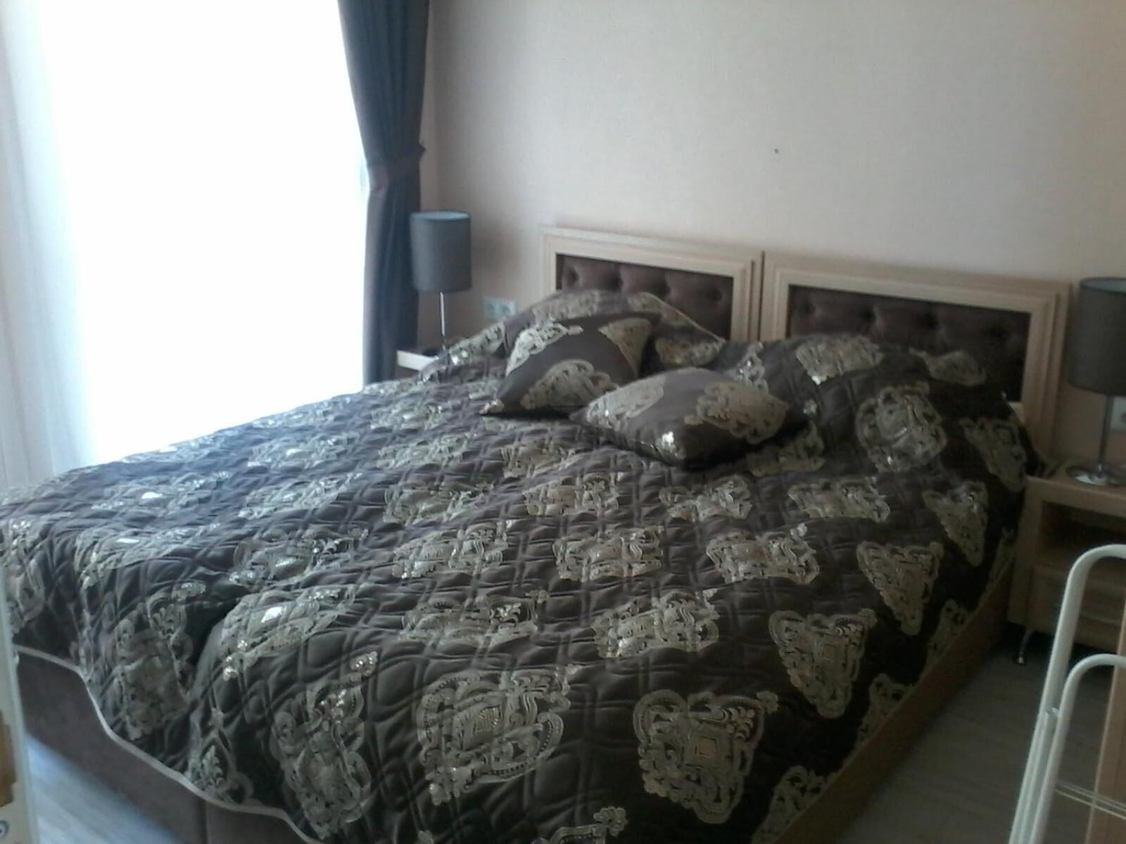 A bed with a patterned bedspread and multiple pillows, a bedside table with a lamp, and a window with curtains.