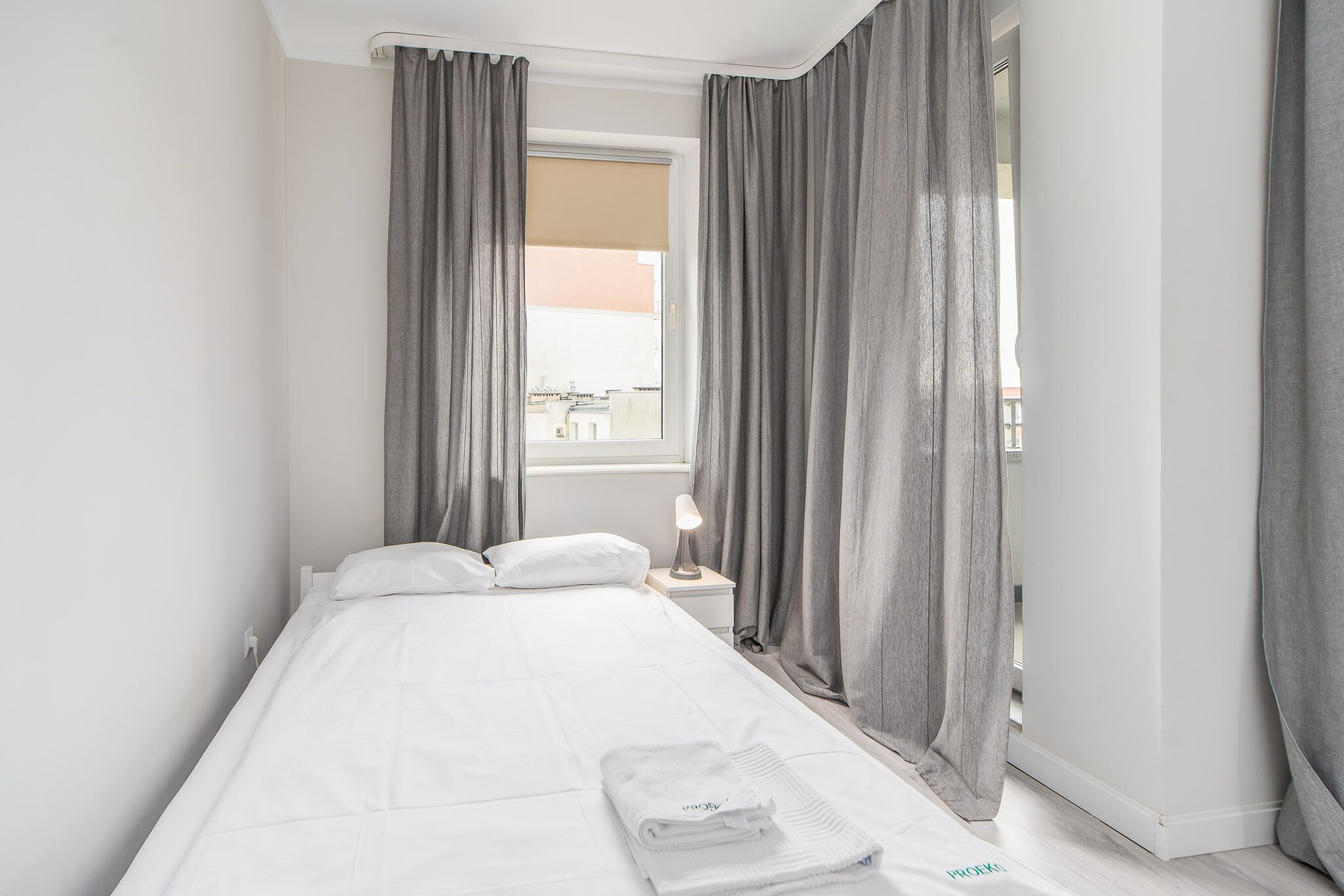 Ein ordentlich bezogenes Bett mit weißen Bettwäsche, ein kleiner Nachttisch mit Lampe und graue Vorhänge, die ein Fenster verdecken.