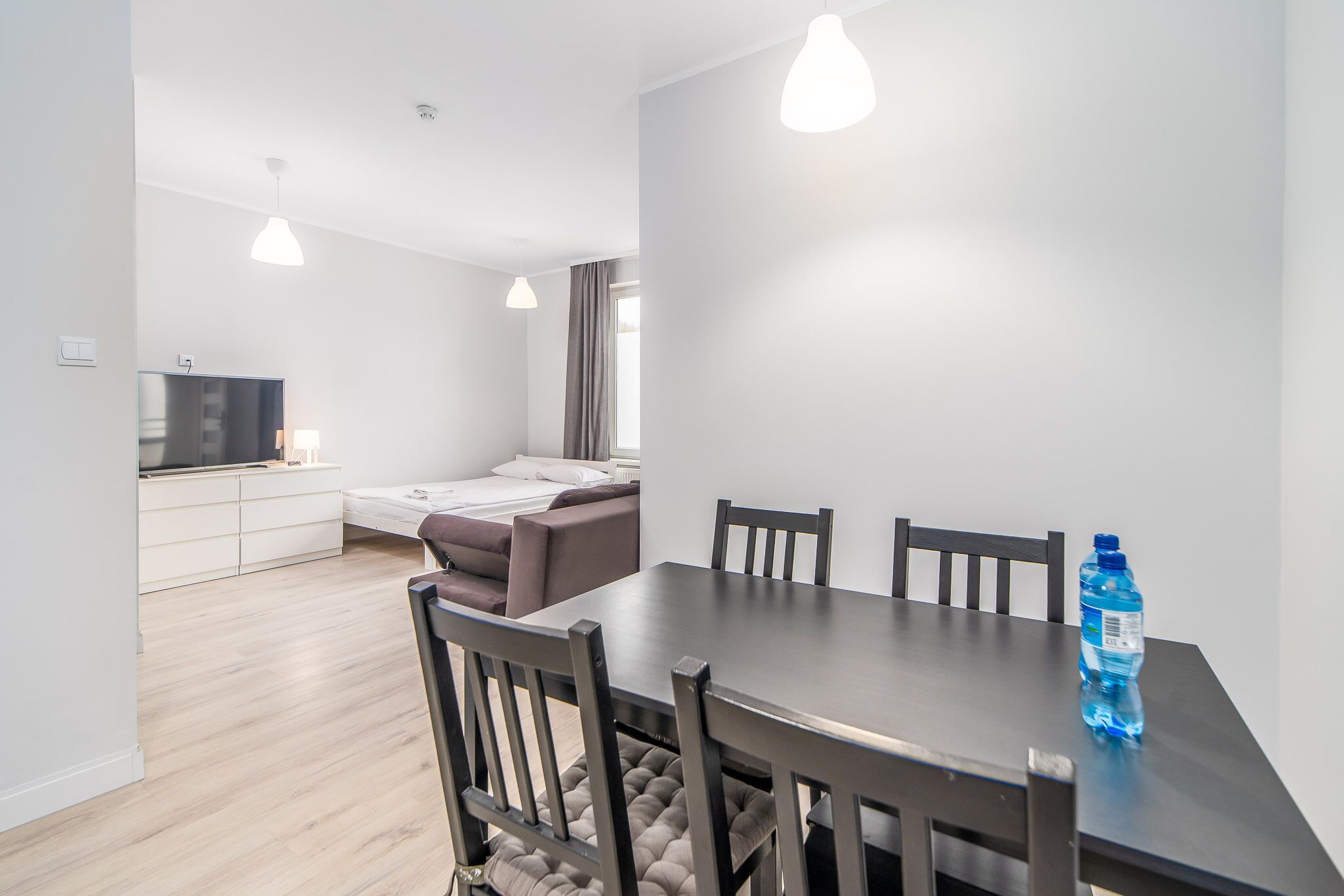 A modern, well-lit room with a dining table, chairs, a sofa, and a TV in the background.